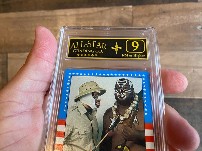 May include: A graded trading card featuring two wrestlers in masks. The card is encased in a clear plastic holder with a gold and black label that reads "ALL-STAR GRADING CO." and a "9" grade. The card has a blue border with white stars.