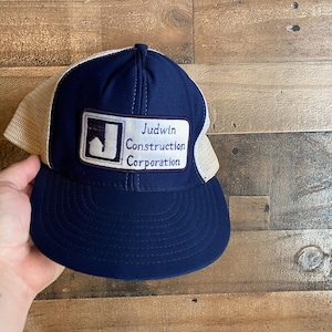 Peut inclure: Un chapeau de camionneur bleu et blanc avec un patch qui indique "Judwin Construction Corporation".