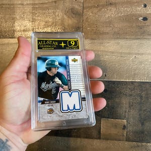 Può includere: Una carta collezionabile di baseball in una custodia di plastica trasparente. La carta presenta un giocatore di baseball in uniforme, con il logo della squadra e il nome "Bobby Crosby". La carta è valutata da "All-Star Grading Co."