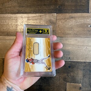 Puede incluir: Una tarjeta de béisbol en un estuche de plástico transparente, calificada por All-Star Grading Co. La tarjeta presenta a un jugador de béisbol con uniforme y el texto "Piece of the Game". La tarjeta tiene un fondo dorado y blanco.