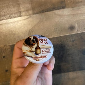 May include: A round button with a white background and a picture of a Saint Bernard dog holding a bottle of Drambuie liqueur. The text on the button reads "Meet Max" and "The Saint Bernard Who Switched to Drambuie Liqueur."