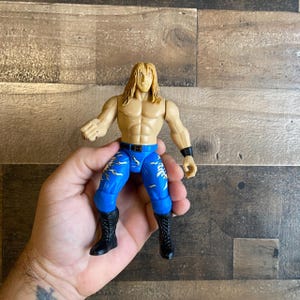 May include: A plastic action figure of a wrestler wearing blue and white patterned pants with black boots. The figure has blonde hair and a muscular build.
