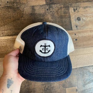 May include: A blue denim trucker hat with a white mesh back. The hat has a white circle patch with a black anchor and the letters "C.C." inside the anchor.