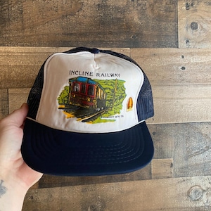 May include: A white and blue trucker hat with a vintage illustration of a red train on the front. The text "INCLINE RAILWAY" is printed in black above the train. The text "LOOKOUT MTN TN" is printed in black below the train.