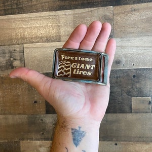 May include: A brown leather belt buckle with a silver metal frame. The buckle features a tire tread design and the text "Firestone Giant Tires".