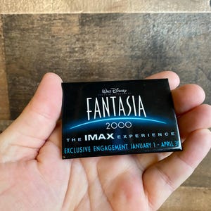 May include: A black and blue button with the text "Walt Disney Pictures Presents Fantasia 2000 The IMAX Experience Exclusive Engagement January 1 - April 30"