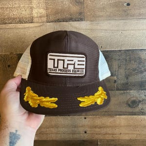 May include: Brown and white trucker hat with a brown patch that says "TPE Texas Process Equip. Co.". The hat has two gold embroidered oak leaves on the brim.