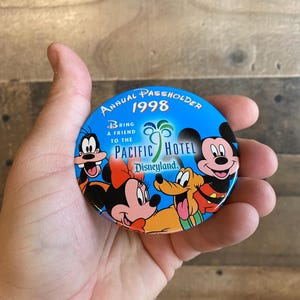 Op de afbeelding: Een blauwe en witte knop met de tekst "Annual Passholder 1998" en "Bring a friend to the Pacific Hotel Disneyland." De knop toont Mickey Mouse, Minnie Mouse, Goofy en Pluto.