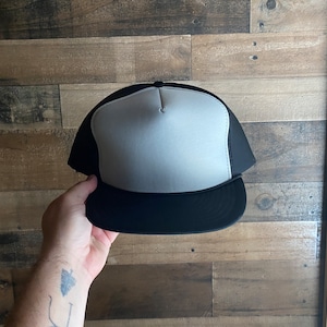 May include: A black and white trucker hat with a mesh back. The front of the hat is white and the brim is black.