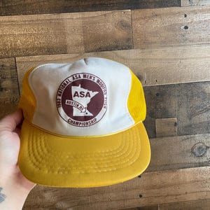 Op de afbeelding: Een witte en gele baseballpet met een donkerrode patch waarop "1983 National ASA Men's Modified Pitch Championship" en een afbeelding van de staat Minnesota staat. De patch bevat ook de tekst "ASA" en "Austin, MN".