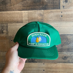 Peut inclure: Casquette de baseball verte avec un patch qui dit "Bendickson Pump & Irrigation" sur fond bleu et deux poires.