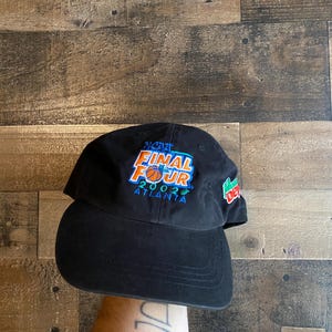 Puede incluir: Gorra de béisbol negra con el texto "NCAA FINAL FOUR 2002 ATLANTA" en azul, naranja y blanco. La gorra tiene una visera curva y un logotipo verde y rojo en el lateral.