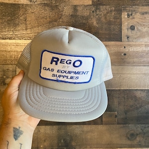 Peut inclure: Un chapeau de camionneur gris et blanc avec un patch bleu et blanc qui indique "REGO BY GAS EQUIPMENT SUPPLIES".