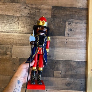 May include: A wooden nutcracker figurine with a black face, red and blue clothing, and a gold crown. The nutcracker is holding an axe and standing on a red base.