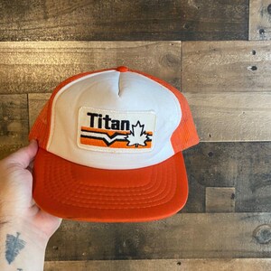 Peut inclure: Casquette de camionneur orange et blanche avec un logo qui dit "Titan" et une feuille d'érable.