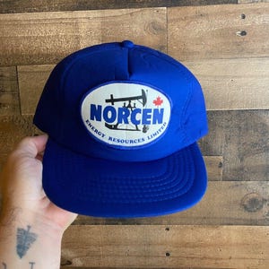 Peut inclure: Un chapeau de camionneur bleu avec un patch blanc représentant une plate-forme pétrolière noire et le texte "Norcen Energy Resources Limited".
