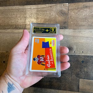 Op de afbeelding: Een beoordeelde sport trading card in een doorzichtige plastic hoes. De kaart toont een American football speler in een rood en wit uniform met nummer 33. De kaart heeft de tekst "Fabric of America" en is beoordeeld met "9" door All-Star Grading Co.