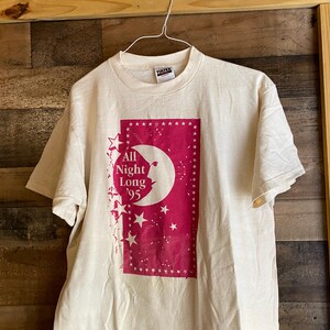May include: A white t-shirt with a pink graphic of a crescent moon with a face and stars. The graphic says "All Night Long '95".