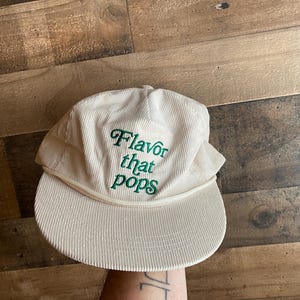May include: A cream-coloured corduroy cap with a curved brim. The cap has the embroidered text "Flavor that pops" in green. The hat has a classic six-panel design and an adjustable strap. The hat is made of corduroy.