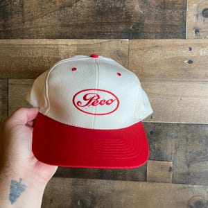 May include: A white and red baseball cap with the word "Peco" embroidered in red script on the front. The cap has a red brim and a red button on top. The hat is made of a sturdy material.