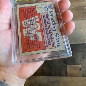 May include: A vintage wrestling trading card featuring Kimchee and Kamala the Ugandan Headhunter. The card is encased in a protective plastic holder. The card's text includes wrestler's names, weight, and origin.