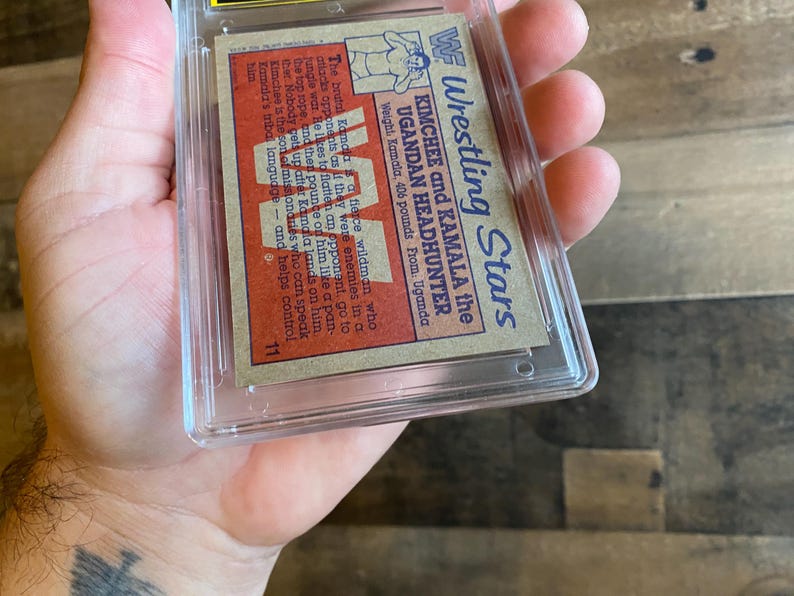 May include: A vintage wrestling trading card featuring Kimchee and Kamala the Ugandan Headhunter. The card is encased in a protective plastic holder. The card's text includes wrestler's names, weight, and origin.