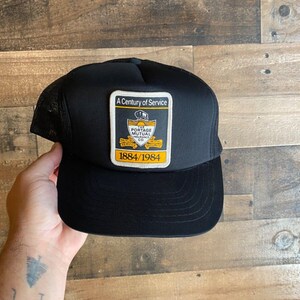 Peut inclure: Casquette de camionneur noire en mesh avec un patch jaune qui indique "A Century of Service" et "The Portage Mutual Insurance Co. 1884/1984".