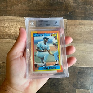 May include: A baseball card featuring Frank Thomas of the Chicago White Sox, graded by Beckett. The card shows Thomas in his White Sox uniform, kneeling on the field. The card is encased in a protective plastic holder.