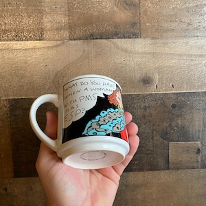 Peut inclure: Une tasse en céramique blanche avec une illustration de bande dessinée en noir et blanc d'une femme aux cheveux roux et une robe à motifs bleus et noirs. Le texte sur la tasse est "What do you have when a woman with PMS has ESP?"