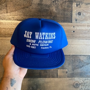 May include: A royal blue trucker hat with white text that reads "Jay Watkins Snow Plowing & Auto Repair 445-4967 Friedens, Pa."