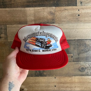May include: Red and white trucker hat with a logo for Intermountain Generator, a company located at 312 W. 5090 S. Murray, Utah. The logo features a cartoon of a man riding a rocket-powered car.