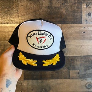 May include: A white and black trucker hat with a black mesh back. The front of the hat has a white oval patch with the text "Tarnes Electric Ltd. Kindersley" in black and red lettering. The hat has two gold embroidered oak leaf designs on the brim.