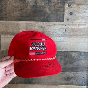 May include: Red baseball cap with a white Jolly Rancher Racing Team logo and a pink braided band. The cap has black signatures on the front.