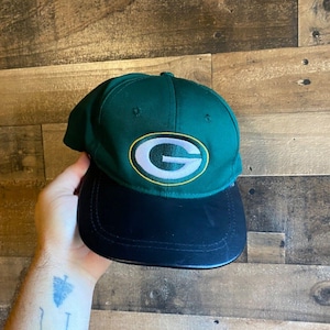 Puede incluir: Gorra de béisbol verde y negra con una "G" bordada en verde y blanco en la parte delantera.