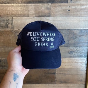 May include: A navy blue trucker hat with a white mesh back. The hat has the text "We live where you spring break" in white lettering with a small flame graphic.