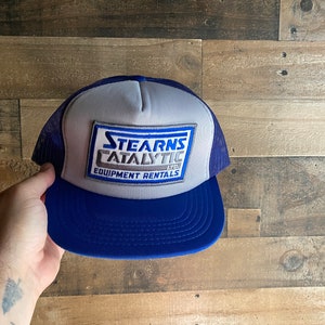 Peut inclure: Un chapeau de camionneur bleu et gris avec un patch blanc qui indique "Stearns Catalytic Ltd. Equipment Rentals".