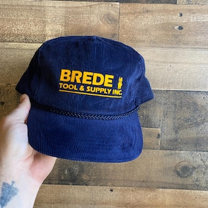 May include: A navy blue corduroy baseball cap with a yellow embroidered logo that reads "Brede Tool & Supply Inc."