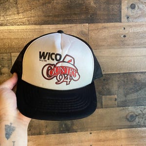 Può includere: Un cappello da camionista bianco e nero con un logo rosso e bianco che dice "WICO 94.3 FM COUNTRY 94".
