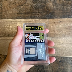 Può includere: Una carta collezionabile sportiva valutata con Taylor Stubblefield, un wide receiver. La carta è racchiusa in un supporto di plastica trasparente con un'etichetta All-Star Grading Co. e un voto di 9. Il testo è "SENIOR STANDOUTS".