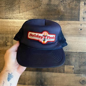 Peut inclure: Un chapeau de camionneur bleu marine avec un patch rouge, blanc et jaune qui dit "Holiday Foods" avec un graphique de ballon à air chaud.