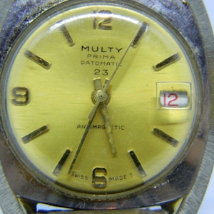 May include: Close-up of a vintage MULTY PRIMA DATOMATIC 23 wristwatch. The watch has a gold-colored face with black markers and hands. The date is displayed at the 3 o'clock position. The watch is marked "SWISS MADE".