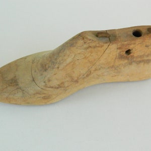 Puede incluir: Horma de zapato de madera de color marrón claro, con punta y talón redondeados. La horma tiene una grieta y dos agujeros cerca del talón. Herramienta utilizada en la fabricación de calzado.