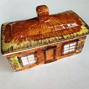 May include: A ceramic cottage-shaped trinket box with a thatched roof and chimney. The box features brown, yellow, and white colors, with two windows and a door. The cottage is decorated with floral accents.