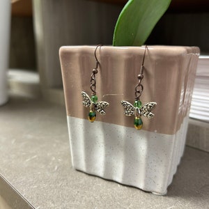 May include: A pair of dangle earrings with a dragonfly design. The dragonfly is made of gold-toned metal with green accents. The earrings are hanging from a brown wire.