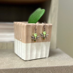 May include: A pair of silver dragonfly earrings with green and black accents. The earrings are hanging from a white and brown ceramic planter.