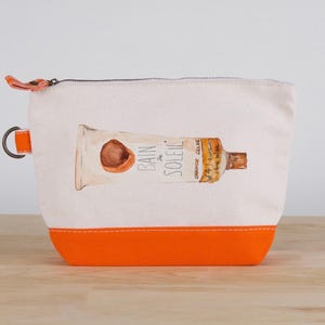 May include: A white canvas cosmetic bag with an orange bottom and zipper pull. The bag features a watercolor illustration of a sunscreen tube with the words "Bain de Soleil" printed on it. A small metal ring is attached to the side.