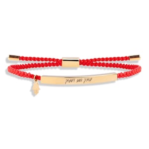 Könnte beinhalten: Rotes Armband mit einem goldenen Barren-Anhänger, der mit hebräischem Text graviert ist, und einem goldenen Hamsa-Anhänger.