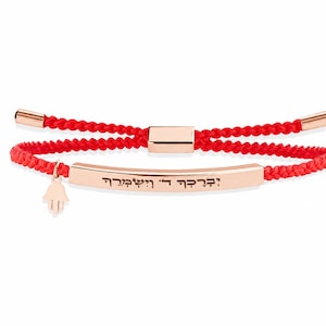 May include: A rose gold-toned bar bracelet featuring a vibrant red woven cord, an adjustable sliding clasp, and a small hamsa hand charm. The central metal bar is engraved with Hebrew text, and the cord ends are finished with rose gold-toned metal caps.