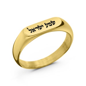 May include: A gold-toned stainless steel ring with a rectangular band engraved with the Hebrew phrase "שלום לך אלה" in black.