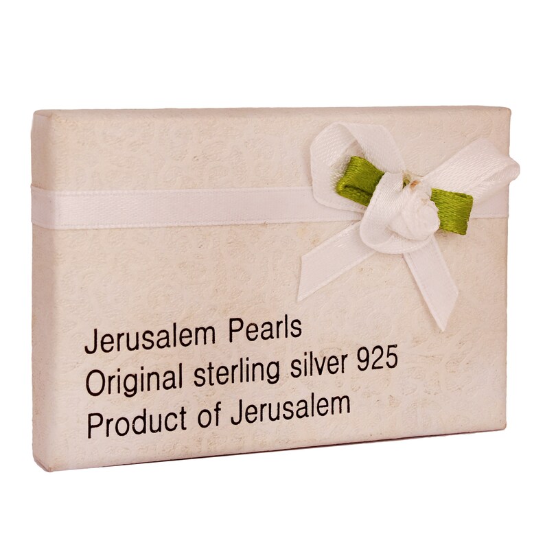 Pu&ograve; includere: Una scatola regalo bianca con un nastro bianco e un fiocco verde. La scatola ha la scritta "Jerusalem Pearls Original sterling silver 925 Product of Jerusalem" stampata su di essa.